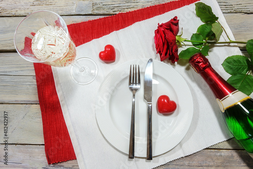 Table setting for a Valentines dinner, red rose and two hearts, chapagne glass and bottle around a white plate on napkins and on a rustic wooden table, love concept, high angle view from above photo