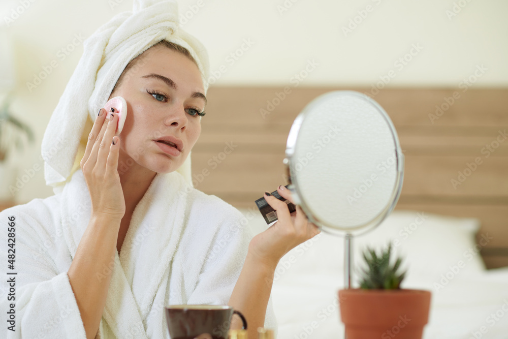 Young woman in bathrobe looking at mirror at her vanity and applying powder foundation Young woman in bathrobe looking at mirror at her vanity and applying powder foundation