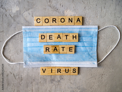 Words CORONAVIRUS DEATH RATE lined with wooden letters on a background of blue surgical mask.