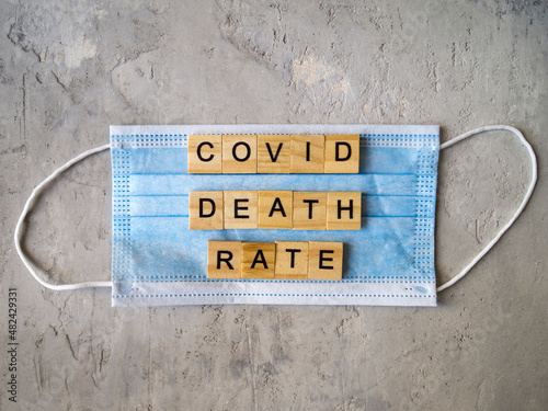 Words COVID DEATH RATE lined with wooden letters on a background of blue surgical mask.