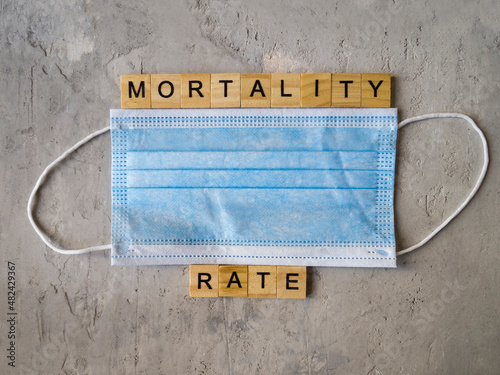 Words MORTALITY RATE lined with wooden letters on a background of blue surgical mask.
