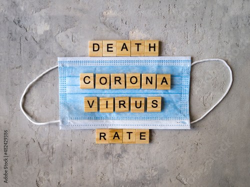 Words DEATH RATE CORONA VIRUS lined with wooden letters on blue surgical masks on a grey background.