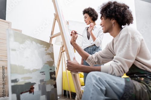 Young african american woman holding paintbrush near canvas and blurred boyfriend painting at home.