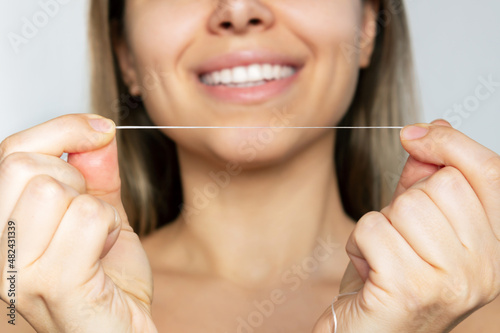 Dental floss in the hands of a young smiling woman in disfocus cleaning her teeth after meal isolated on a gray background. Dental health care, oral hygiene, evening routine. Dentistry concept