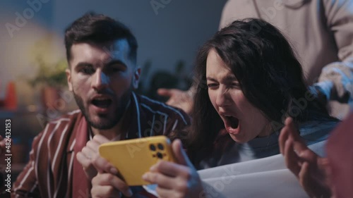 Positive cheerful people looking at the smartphone with happy emotions during football game. Excited football fans using modern smartphone on at home. Football fans concept.