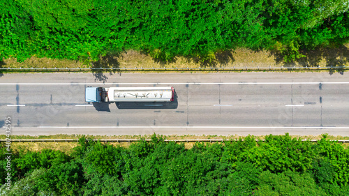Aerial. Gasoline fuel truck driving by the cracked highway road between green forest. Top view from drone.