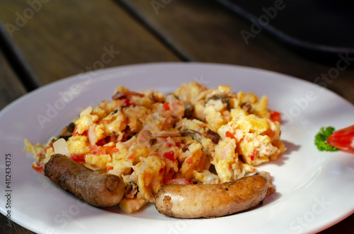 a plate of scrambled eggs with ham, bacon and sausages, and various vegetables, on a table.