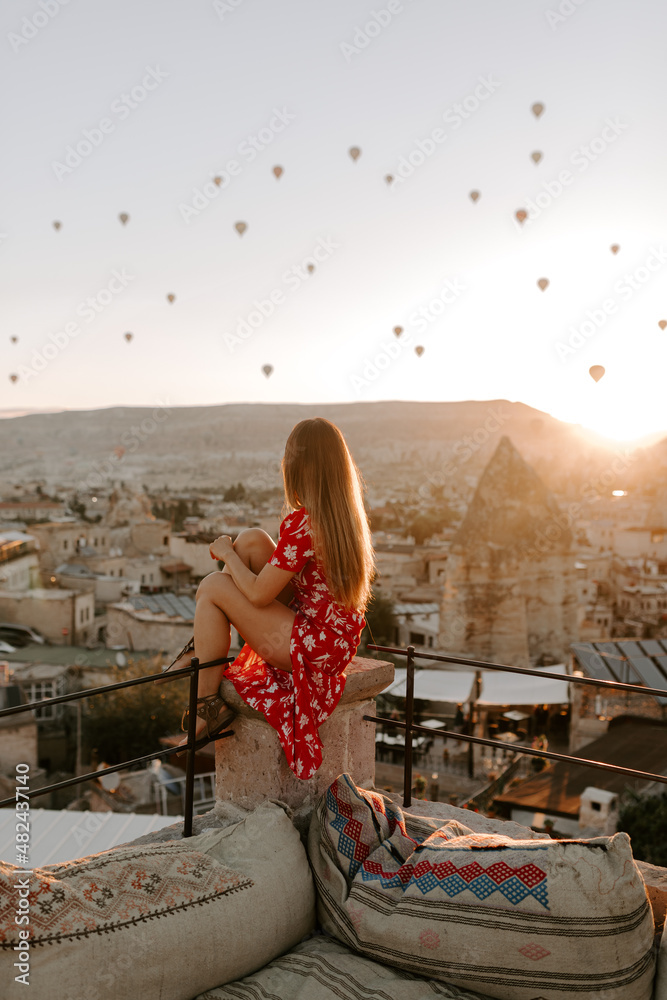 A young female traveller in a red dress is watching hot air balloons in Cappadocia, Turkey at sunrise on a hot summer day
