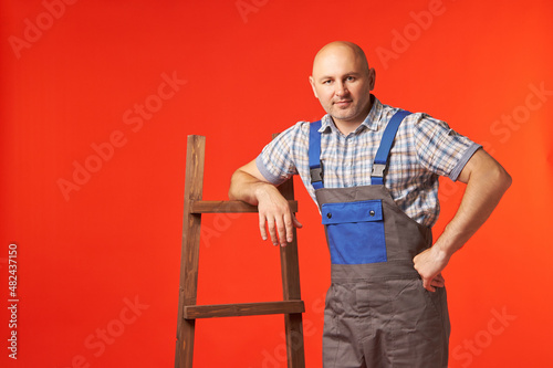 Bald man in work clothes is standing with his hand on the stairs, a photo on a red background. An employee is resting, holding onto a wooden ladder. © Aleksandr