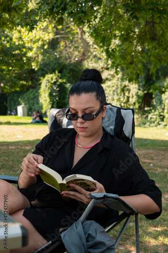 Girl in sun glasses reading book in beautiful weather at outside. She is sitting on chair. Green park. Vertical photo.