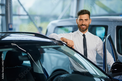 Portrait of handsome bearded man in formal clothes posing near modern luxury car at showroom. Successful businessman buying new vehicle at shop. © serhiibobyk