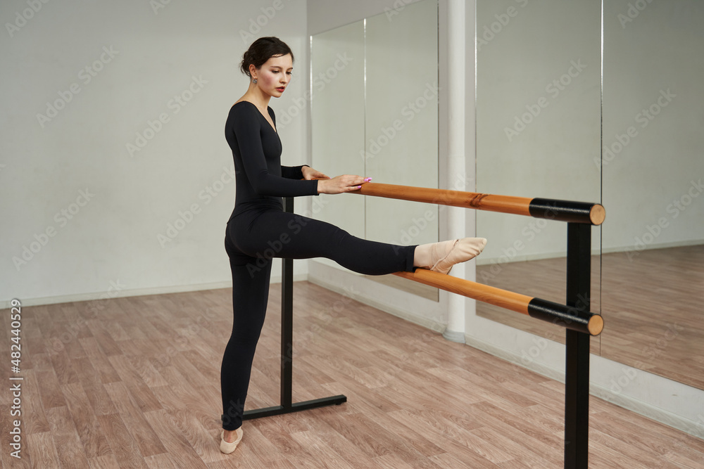a ballerina in a black tight-fitting suit put her foot in pointe on a ballet barre in front of a mirror