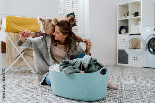 Little girl wants to play with mom who is busy with chores woman sits on bathroom floor laundry room sorting clothes for washing grabs her daughter standing behind her back by hands photo