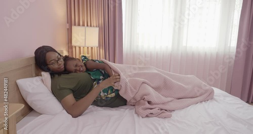 Loving African American mom hugging soothing an adorable, sweet baby girl lying in bed, cheerful family small kid girl and mom embrace cuddle laugh relax in the bedroom.