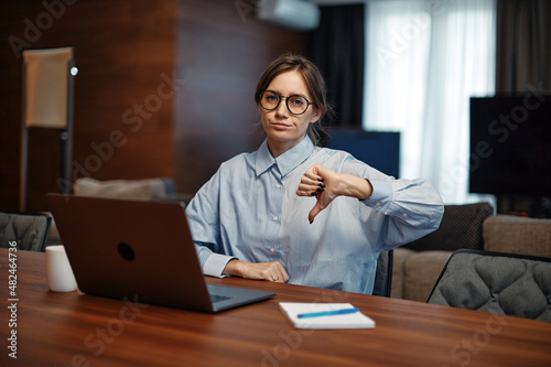 Upset business woman in glasses sit at laptop, showing dislike with thumb down, negative feedback, rejection concept