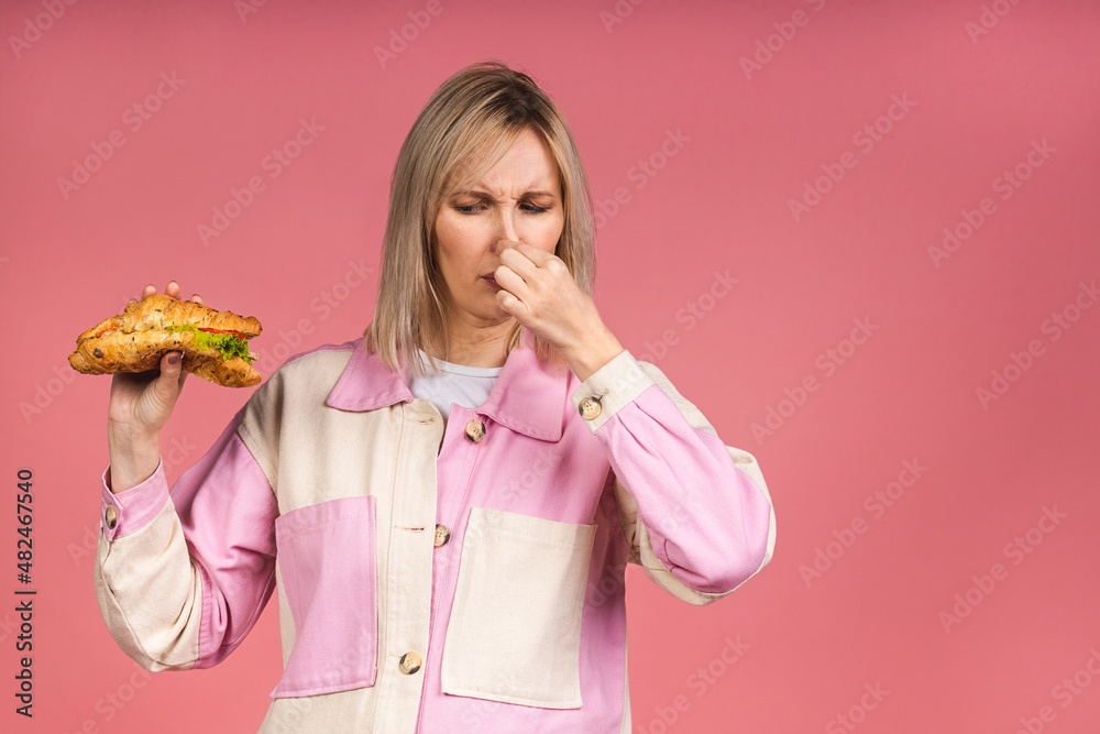 Portrait of a young woman holding her nose because of a bad smell isolated over pink background. Smelling junk food stinky and disgusting, intolerable smell, holding breath with fingers on nose.