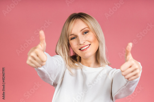 Portrait of a young beautiful cute cheerful blonde woman smiling looking at the camera isolated over pink background. Thumb up sign. © denis_vermenko