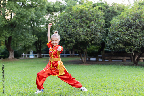 Cute little caucasian girl seven years old in red sport wushu uniform exercising in park at summer day. Lifestyle portrait of kung fu fighter child athlete photo