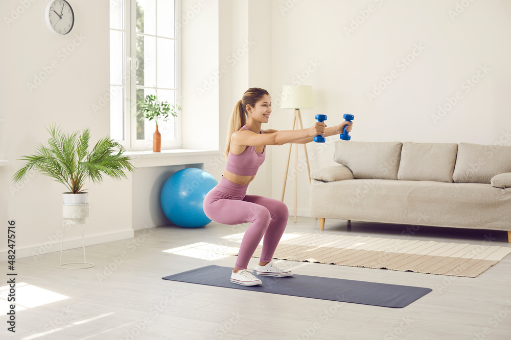 Fototapeta premium Beautiful and slender young woman performs sports exercises at home using dumbbells. Smiling girl in comfortable sportswear sitting down slightly and stretching her arms forward holding two dumbbells.