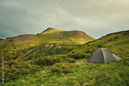 Summer view of small comfortable modern tourist tent on grassy hill under beautiful blue sky on foggy mountains covered with forest background.