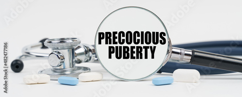 On a white surface are pills, a stethoscope and a magnifying glass inside which is written - Precocious puberty photo