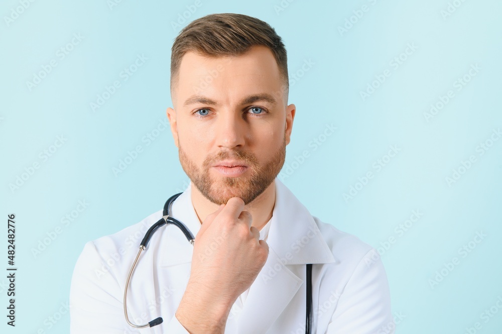 Portrait young bearded doctor with stethoscope over neck in medical coat standing against isolated blue background.