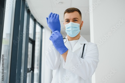Young smiling Caucasian male doctor in white uniform, with eyeglasses and stethoscope around neck putting rubber gloves before patient examination.