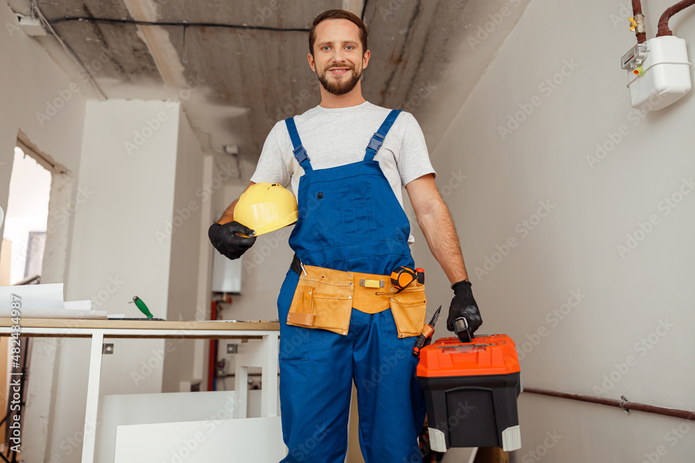 Hard working contractor, handyman in uniform with toolbox smiling at camera while standing indoors Hard working contractor, handyman in uniform with toolbox smiling at camera while standing indoors