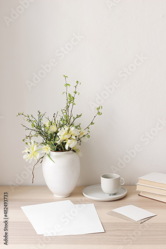 Fototapeta Naklejka Na Ścianę i Meble -  Floral still life. Easter bouquet. Wite, yellow tulips, daffodils flowers. Green birch tree, blueberry branches in ceramic vase pot on wooden table. Vertical. Blank paper cards mockups, cup of coffee