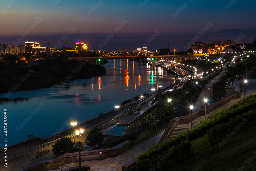 Fototapeta premium Beautiful view of the illuminated Embankment of Tura River and the Chelyuskintsev Bridge at dusk, Tyumen, Russia 