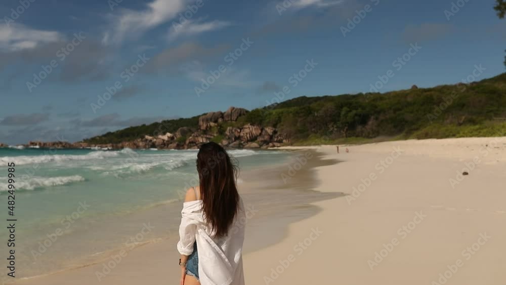 A young female traveller is walking, running on the white sand beach in the tropical island of Seychelles, surrounded by palm trees, waves and turquoise waters A young female traveller is walking, running on the white sand beach in the tropical island of Seychelles, surrounded by palm trees, waves and turquoise waters