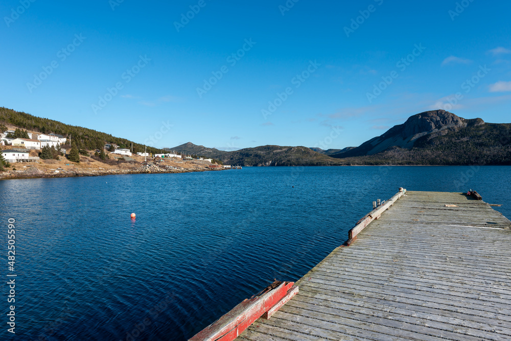 An old wooden pier with red color mooring deck. The wharf juts out into the calm ocean with white community houses and tree covered mountains in the bay or cove. The sky and water are clear blues. 