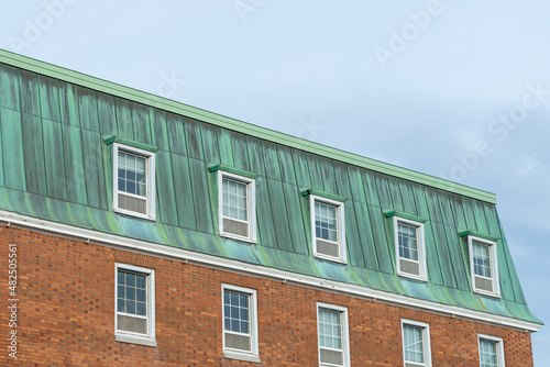 The corner view of a vintage brick building with a copper hip roof that has oxidized to a pale green outer layer, patina.  The bricks are small in a horizontal pattern. There are multiple windows.  photo