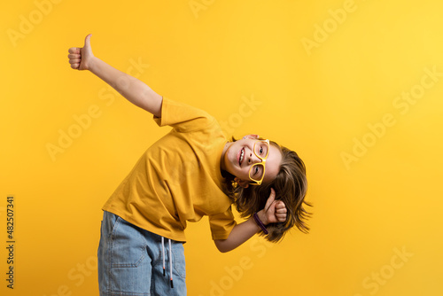 Determined Little Girl 8-10 Years Old in Basic Yellow T-shirt Eyeglasses on Daffodil Color Trend Wall Background Studio. Showing Ok Sign with Fingers. Happy Friendly Looking Child Pointing Thumbs Up