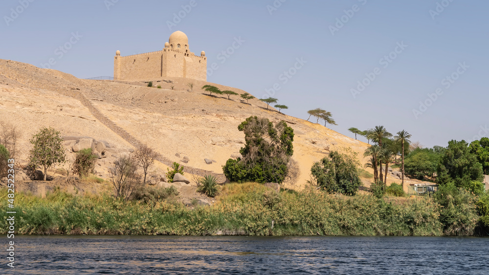 There is green vegetation on the banks of the Nile. At the top of a sand dune, against the blue sky, the ancient mausoleum of the Aga Khan is visible. Egypt. Aswan