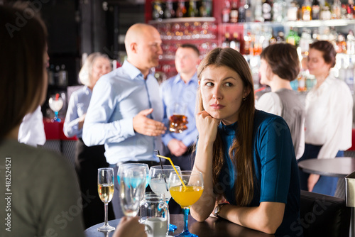 Upset young woman sitting alone at corporate bar party on background with cheerful colleagues..