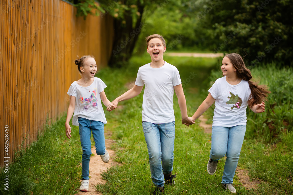 Fototapeta premium Cute little kids are running together on the road. Two sisters and a brother are holding hands, they are having fun, they are happy. The concept of childhood and friendship.