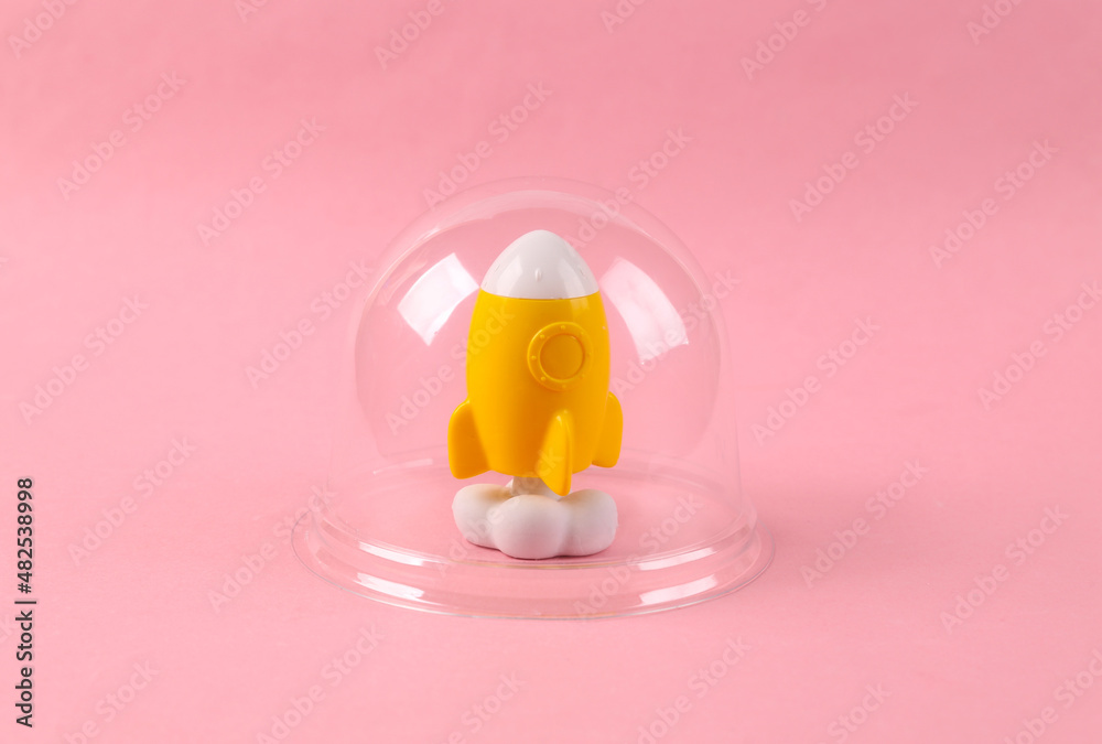 Fun space rocket model under transparent dome on pink background. Protection, isolation concept. Minimal layout Fun space rocket model under transparent dome on pink background. Protection, isolation concept. Minimal layout