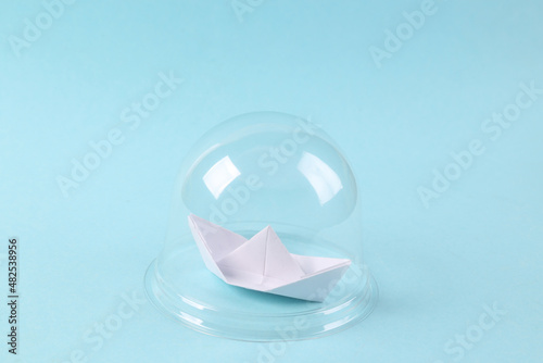 Paper sailboat under transparent dome on blue background. Protection, isolation concept. Minimal layout