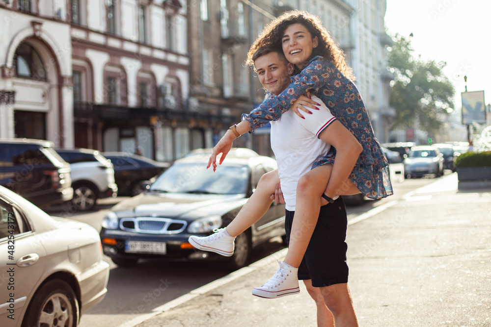 Couple in love. Man carrying girl on his back in the street. Smiling man with beautiful young woman, ride piggyback, having fun together. Relationship concept.