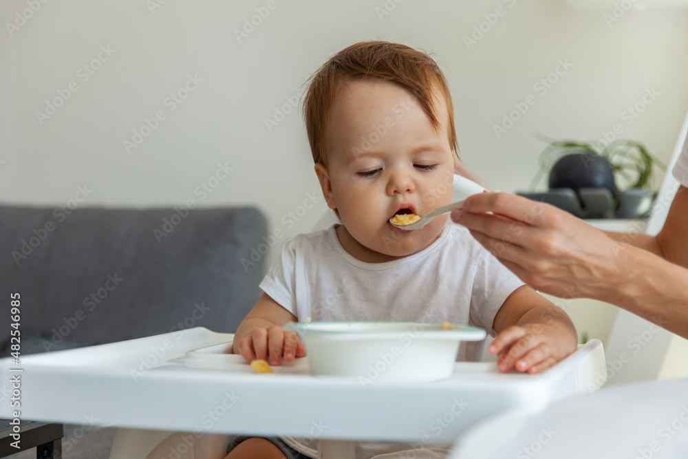 Mom feeds Little child sitting at the children's table at home Mom feeds Little child sitting at the children's table at home