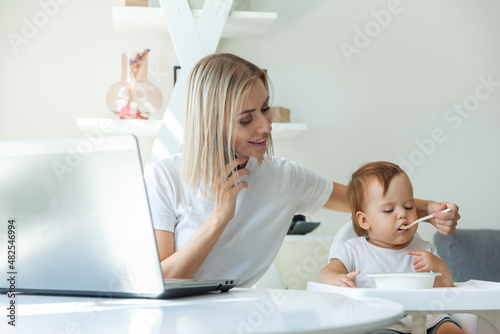 Work from home. Young mother is talking on phone while sitting at table with a laptop and feeding her little daughter. Motherhood, maternity, childhood and care concept.