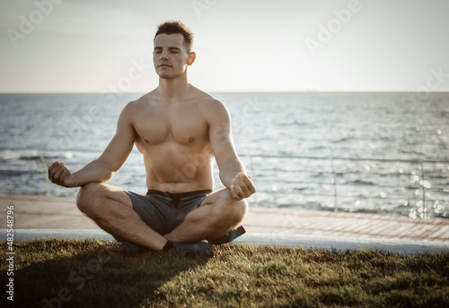 Young muscular man with naked torso meditates in lotus pose at sunrise on beach