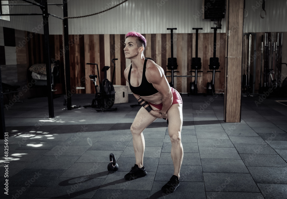 Muscular extraordinary female athlete with short pink hair trains swing with heavy kettlebells. Functional, cross training in modern gym.