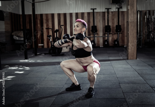 Muscular extraordinary female athlete with short pink hair trains squats with heavy kettlebells. Functional  cross training in modern gym.