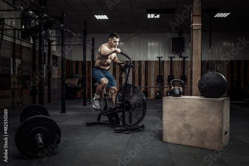 Powerful muscular male athlete with a naked torso trains with an air bike in a modern health club. Functional, cross fit training. Cardio exercise. Healthy lifestyle concept