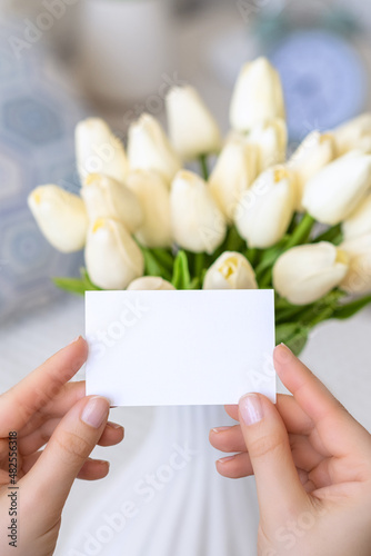 Woman pulling blank greeting card from bouquet of white tulips flowers. Birthday, Copy space. Mock up