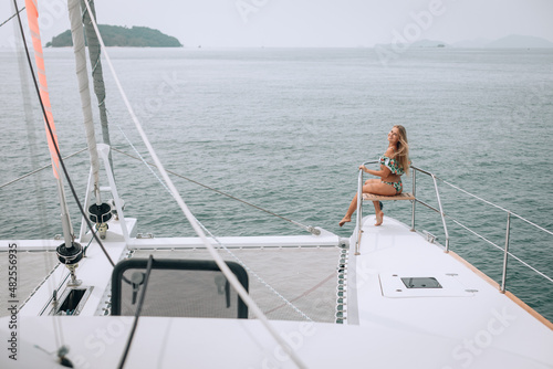 Young blonde smiling girl sits posing on the edge of the deck of a white yacht in a bright colored swimsuit. Background of the open sea with a green island. The concept of tourism and recreation.