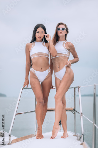 Two attractive young bikini models standing on a deck of expensive yacht. Beautiful blonde and  brunette. A cheeky look at the camera. Background of turquoise big ocean.