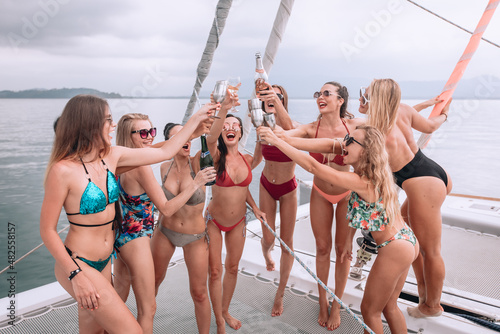 ..Photo from an outdoor resort. Relaxing females ' entertaintment on the open big sea. Cool girls in swimsuits and sunglasses on white luxury yacht drinking champagne for toast, raising her hands.